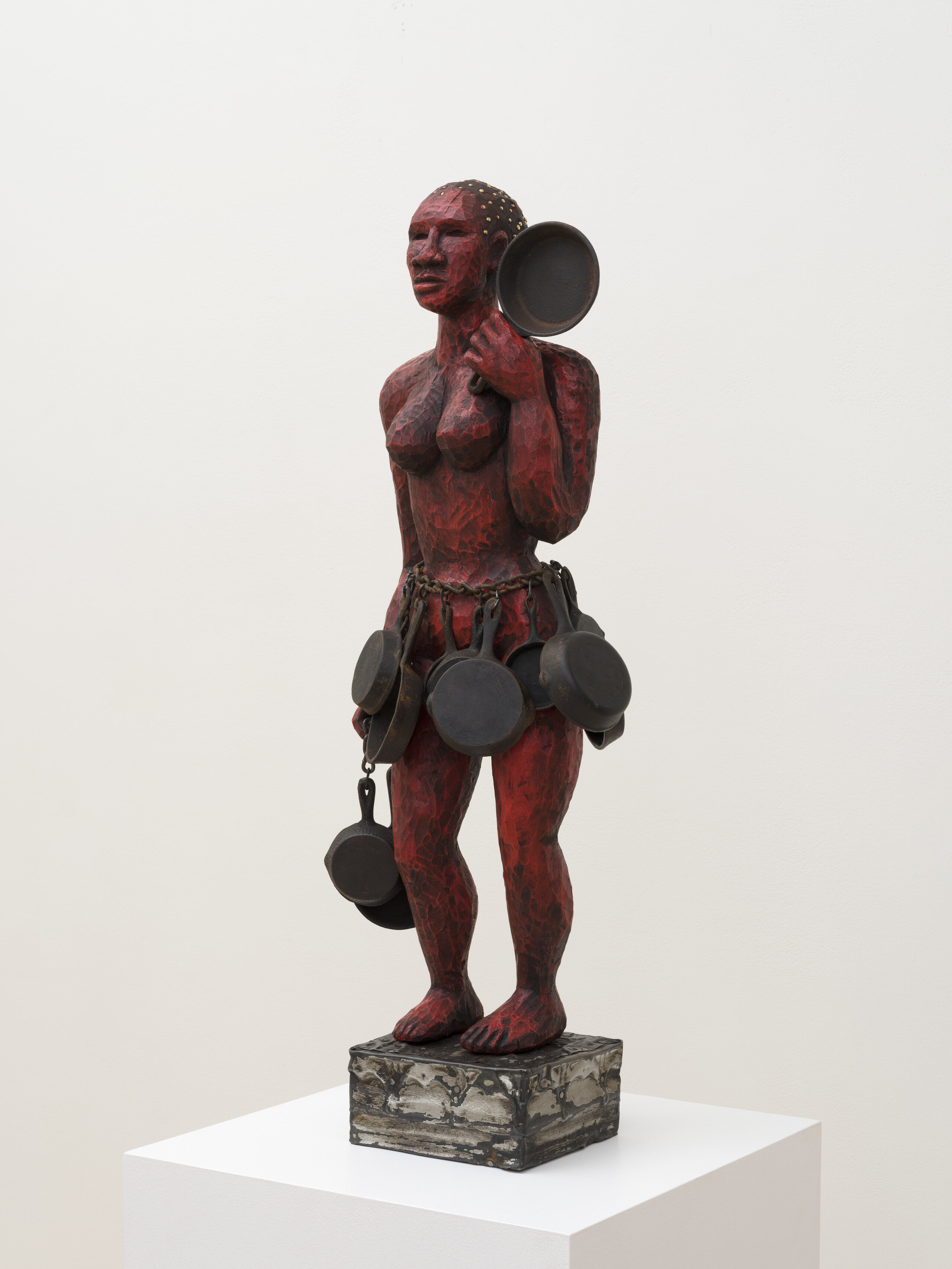 A wooden sculpture is painted red and shaped as a nude woman holding a cast iron skillet in her left hand. She has a heavy chain around her waist with multiple skillets hanging from it like a skirt. She stands on a small wooden platform.