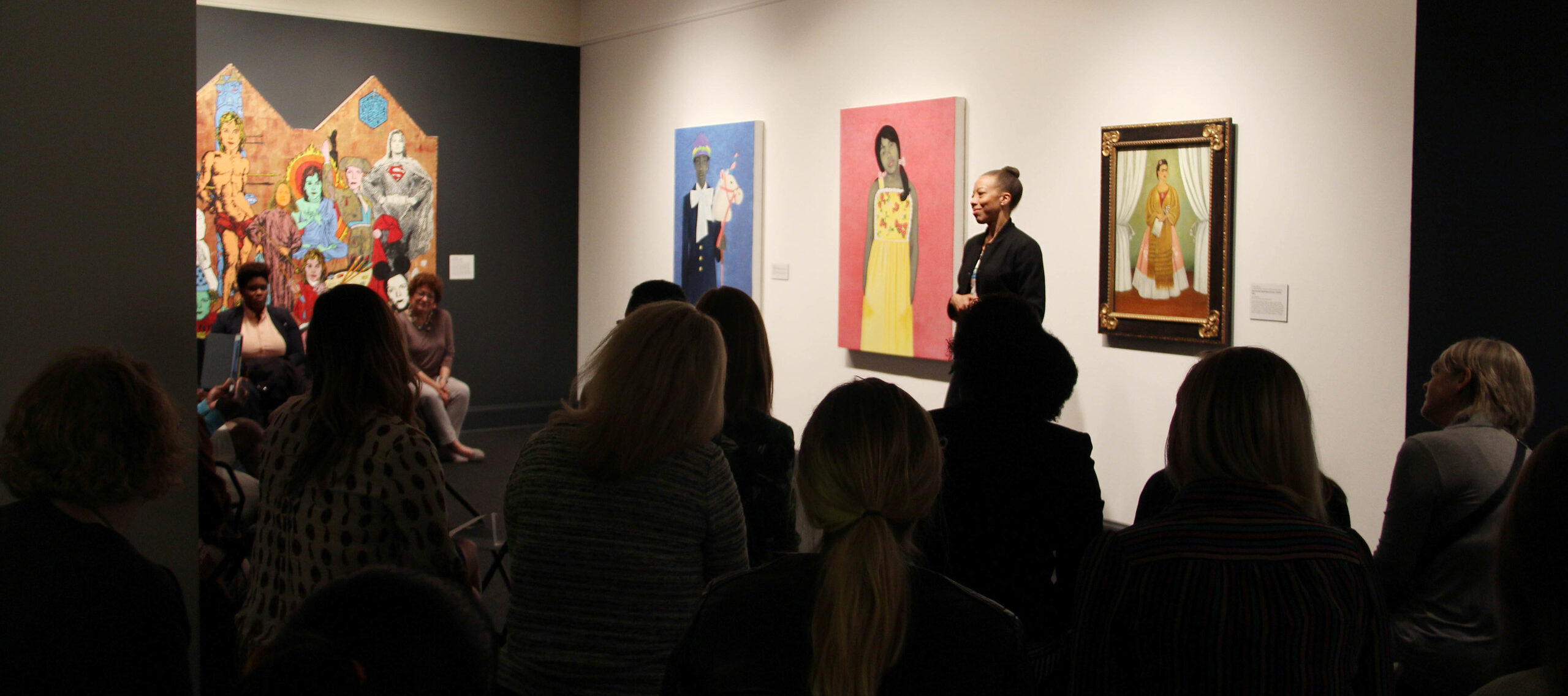 In a museum gallery, an artist with medium skin tone discusses her works to an audience.
