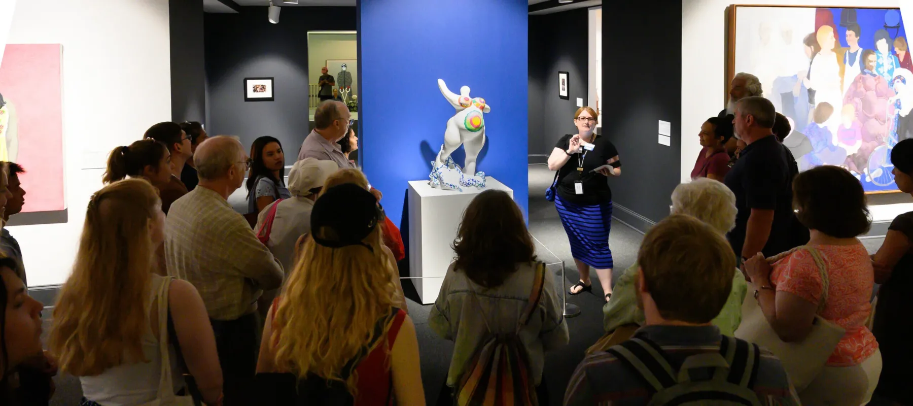 A light-skinned museum docent discusses a white marble sculpture of a female figure covered in bright patterns with visitors in the gallery.