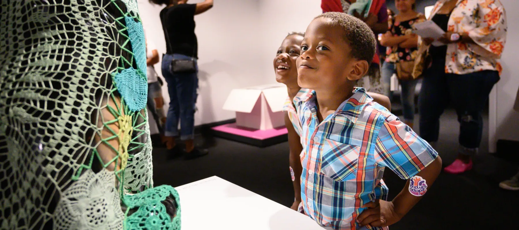 Two young children with medium skin tone stand in front of a dog sculpture covered with green crochet panels. One child stands with hands on hips, eyes turned to look directly at the viewer, and gives a slight smirk.