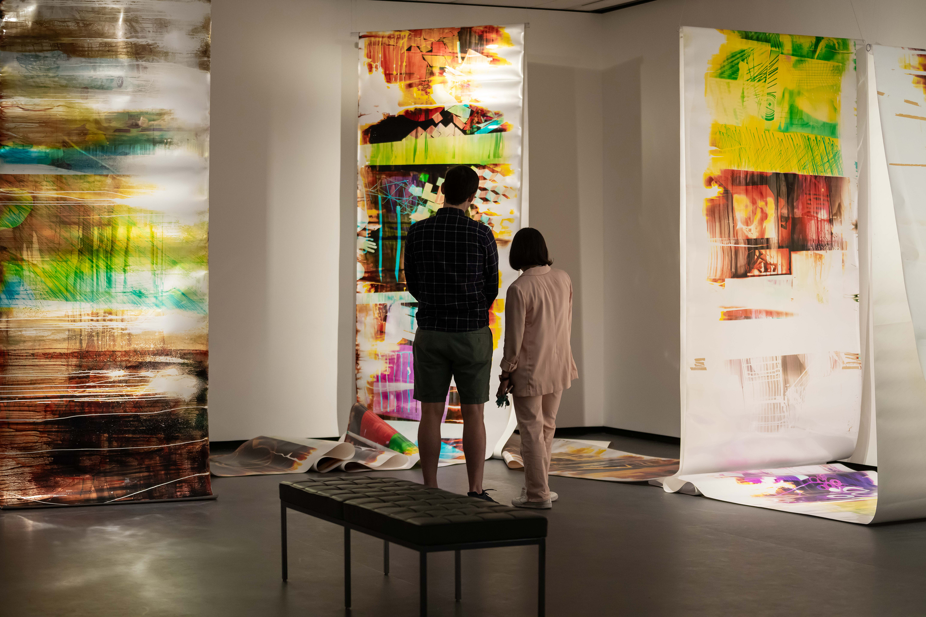 Two museum visitors are standing in front of an artwork. This large-scale, scrolling, colorful, abstract photograph is hung from the ceiling throughout the room.