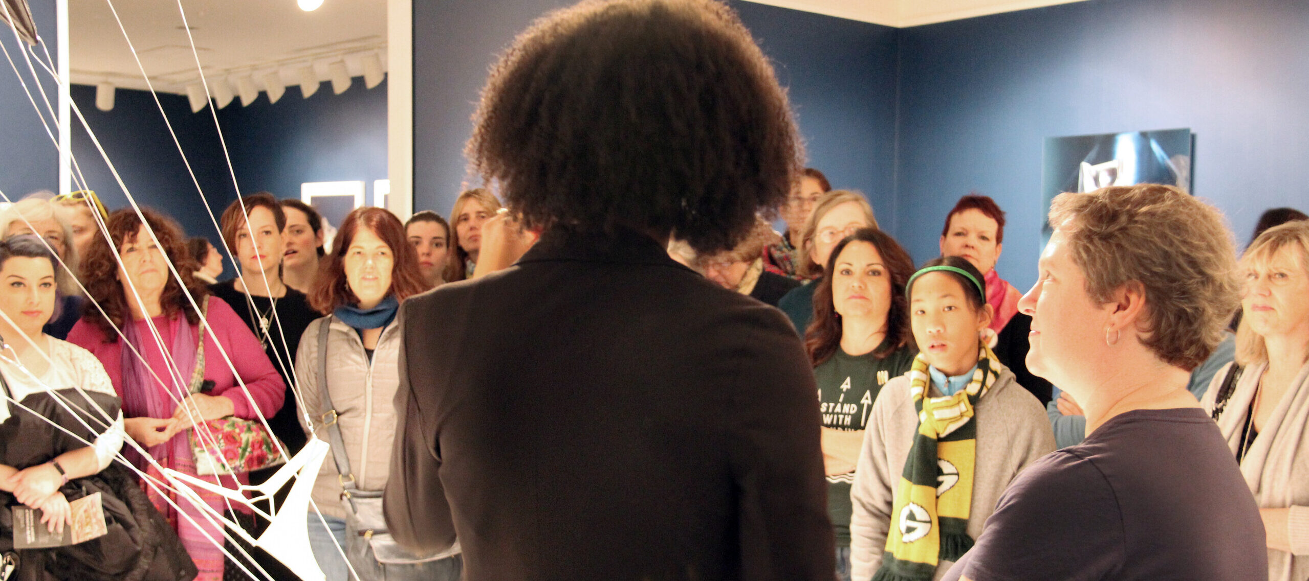 NMWA educator with dark curly hair and a black blazer stands in the foreground with her back to camera as she speaks to a large crowd of visitors about a sculpture composed of hanging strings and fabric in the galleries.