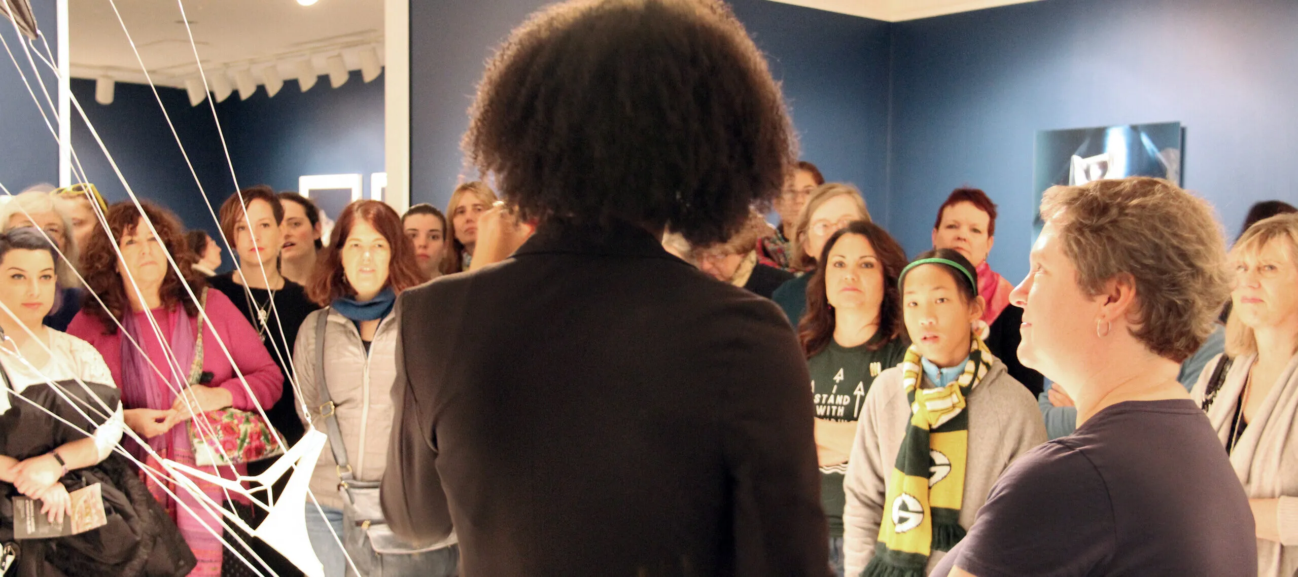 NMWA educator with dark curly hair and a black blazer stands in the foreground with her back to camera as she speaks to a large crowd of visitors about a sculpture composed of hanging strings and fabric in the galleries.