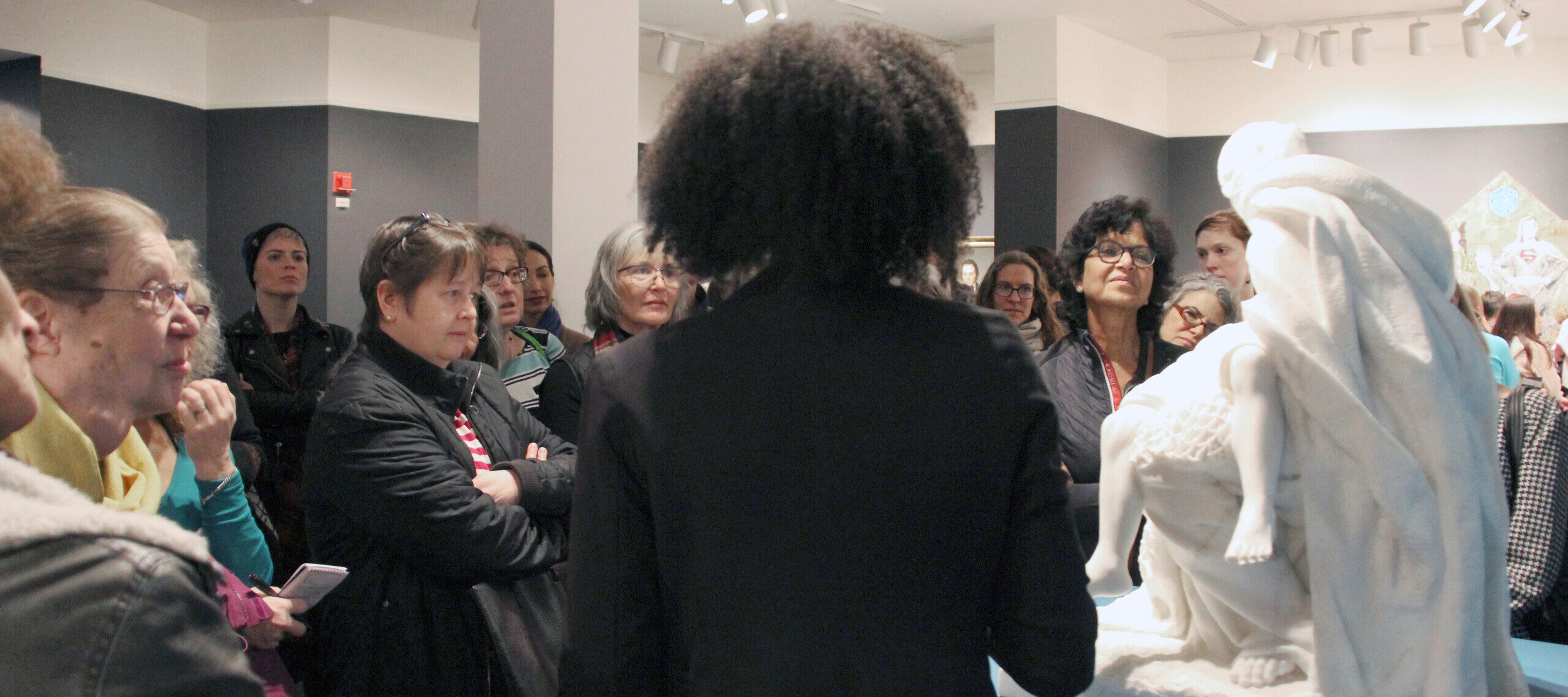 NMWA educator with dark curly hair speaks to a large crowd of visitors about a large marble sculpture.