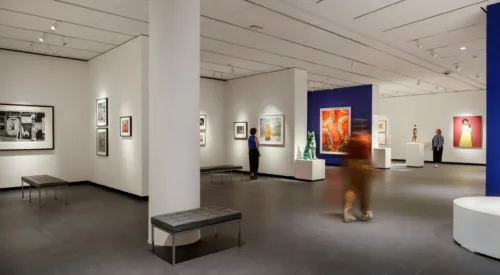 A modern museum gallery is photographed at a wide angle. It features several inset bays in which art of various sizes and mediums is hung. Visitors walk through the galleries and observe the works.