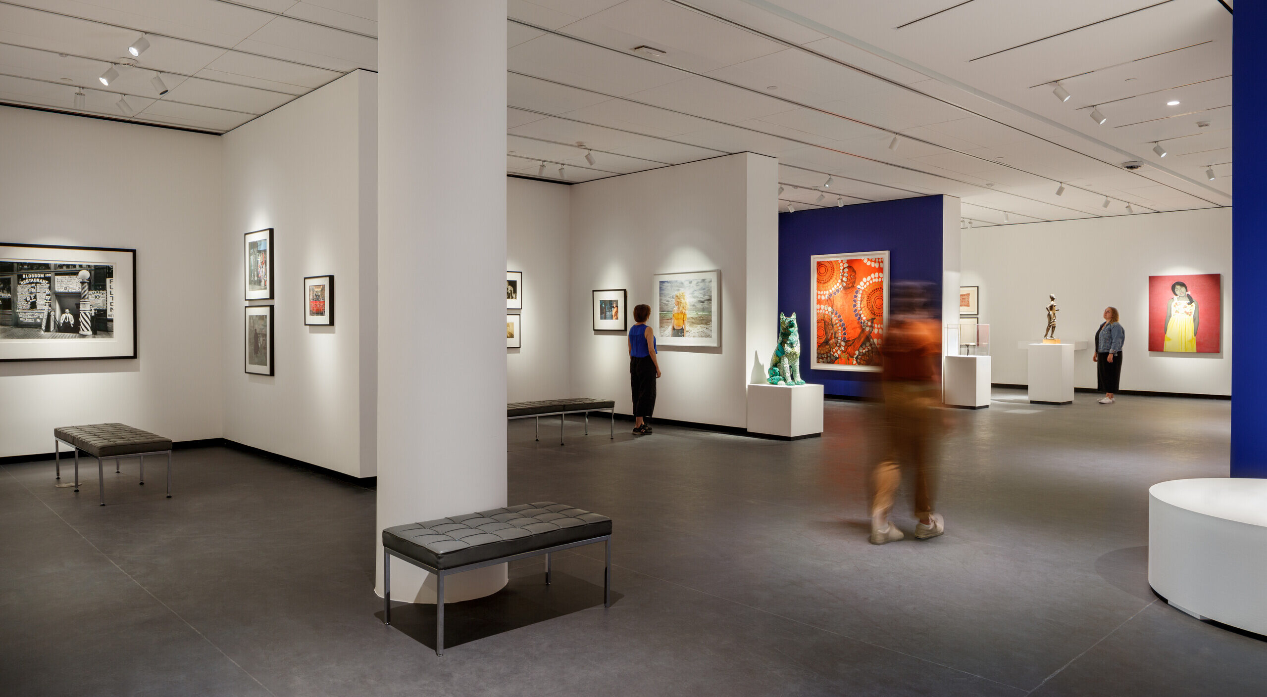 A modern museum gallery is photographed at a wide angle. It features several inset bays in which art of various sizes and mediums is hung. Visitors walk through the galleries and observe the works.