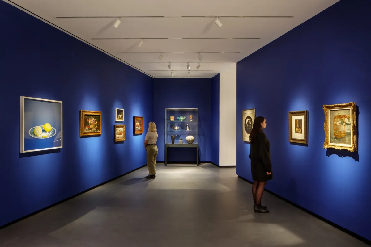 Museum visitors observe artworks hanging on the royal blue walls of a small gallery bay. Paintings of various sizes are on display, and small sculptures are in a glass case.