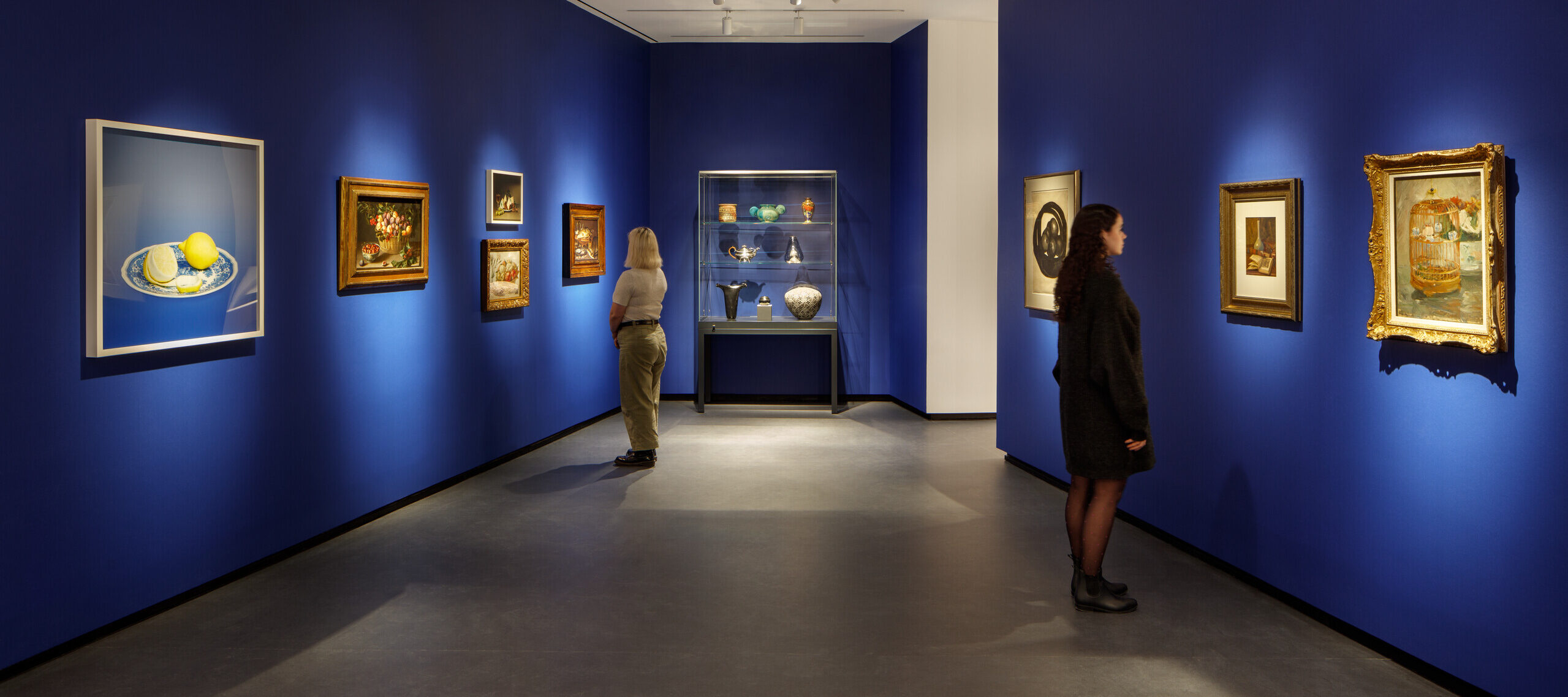 Museum visitors observe artworks hanging on the royal blue walls of a small gallery bay. Paintings of various sizes are on display, and small sculptures are in a glass case.