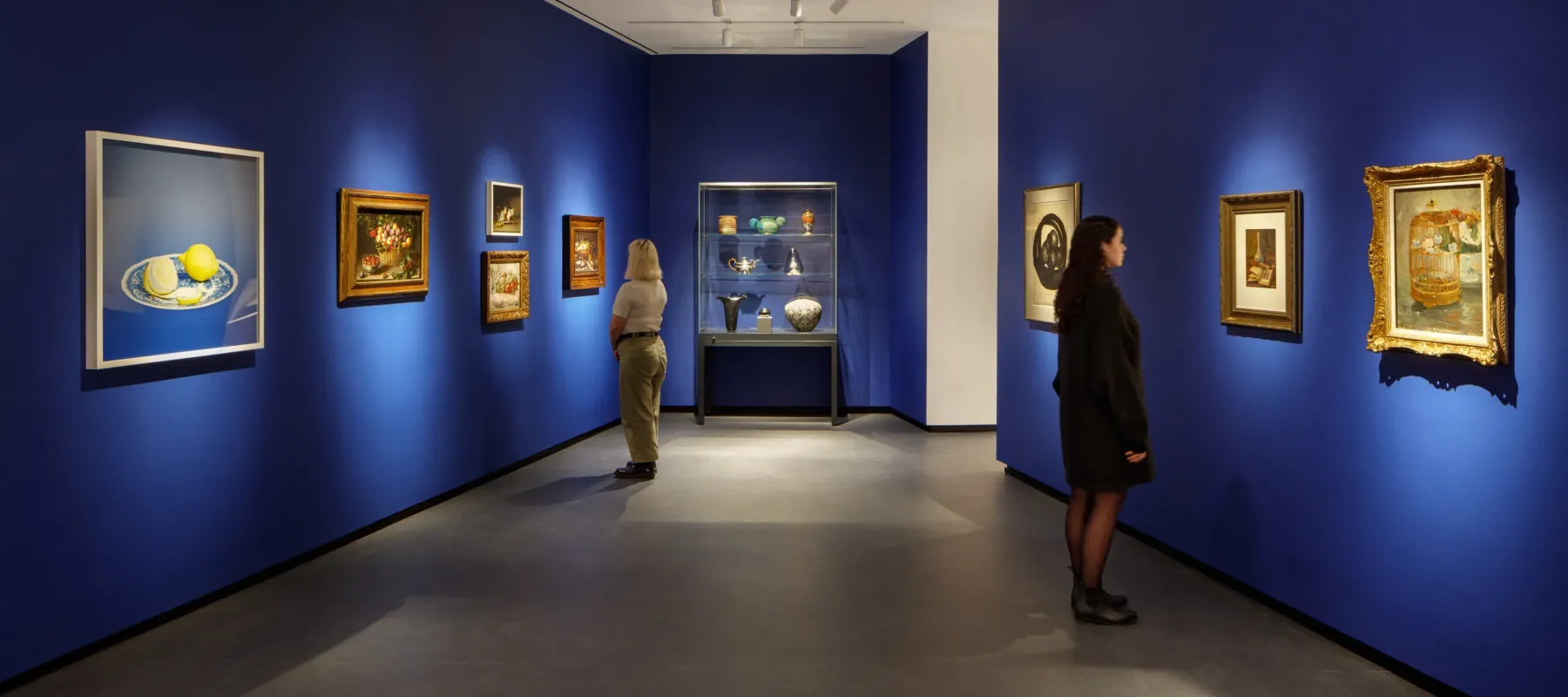 Museum visitors observe artworks hanging on the royal blue walls of a small gallery bay. Paintings of various sizes are on display, and small sculptures are in a glass case.