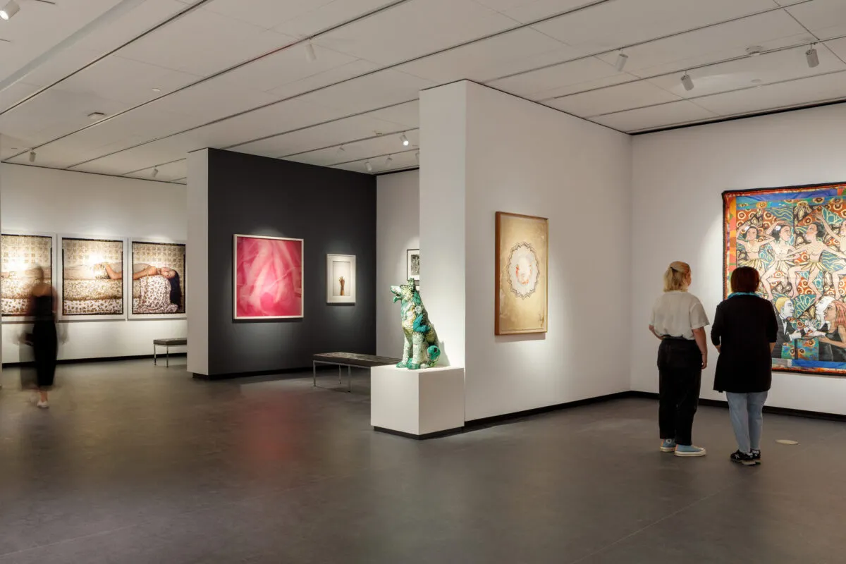 A modern museum gallery is photographed at a wide angle. It features several inset bays in which art of various sizes and mediums is hung. Visitors walk through the galleries and observe the works.