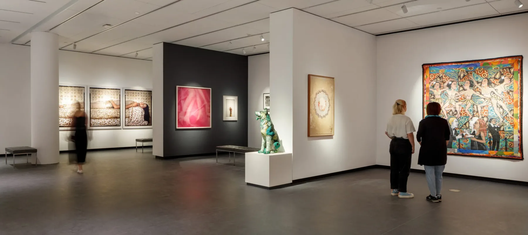 A modern museum gallery is photographed at a wide angle. It features several inset bays in which art of various sizes and mediums is hung. Visitors walk through the galleries and observe the works.