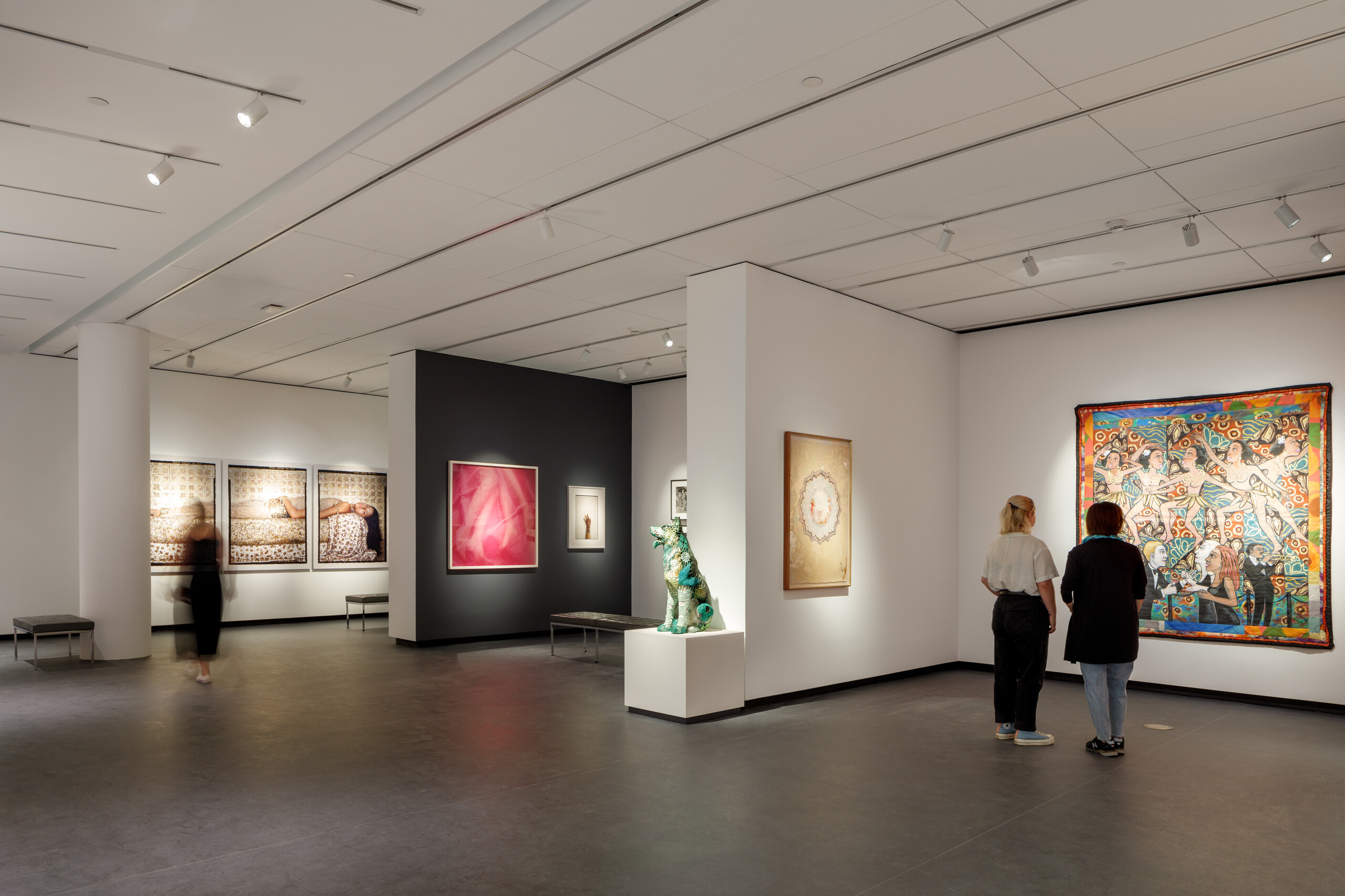 A modern museum gallery is photographed at a wide angle. It features several inset bays in which art of various sizes and mediums is hung. Visitors walk through the galleries and observe the works.