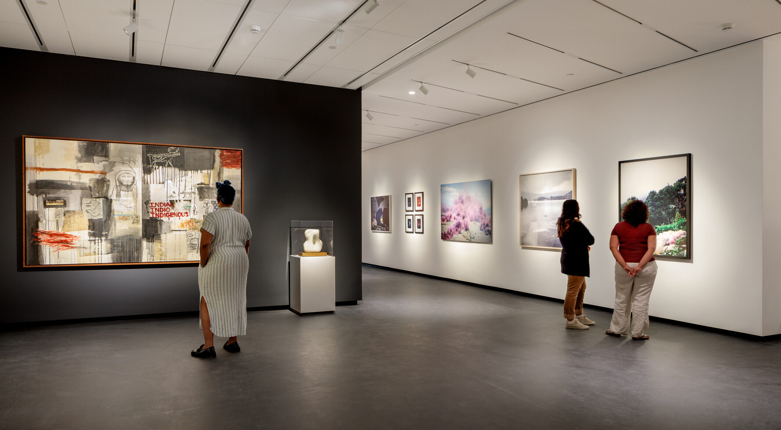 A modern museum gallery is photographed at a wide angle. Visitors observe large artworks on the walls.