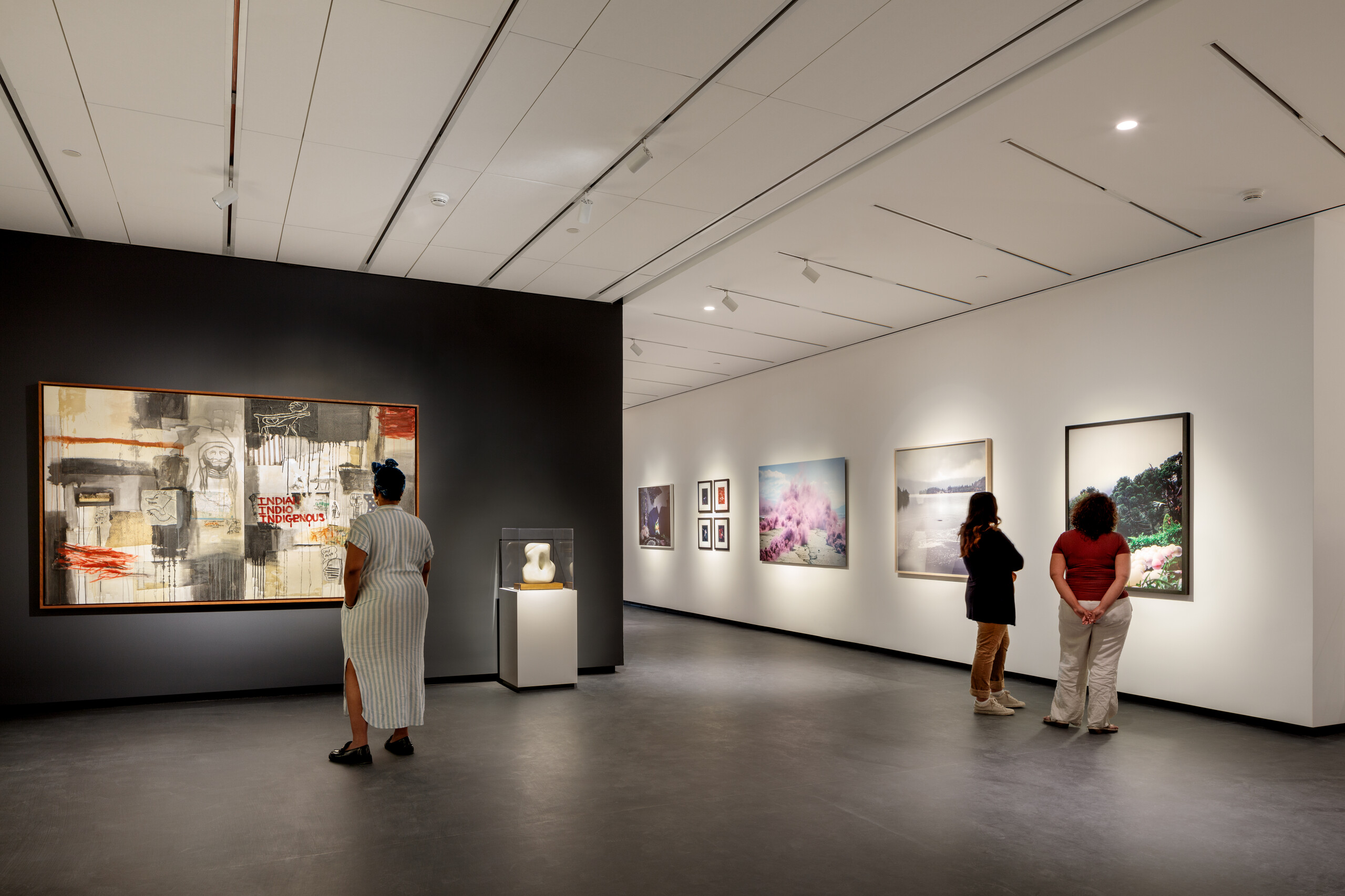 A modern museum gallery is photographed at a wide angle. Visitors observe large artworks on the walls.