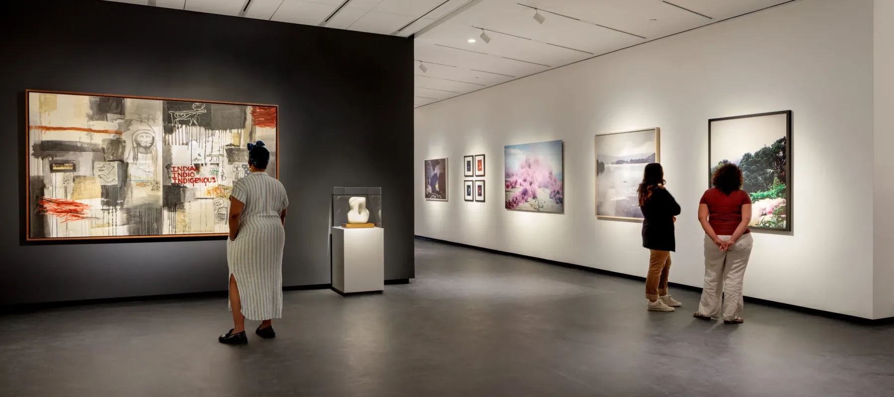 A modern museum gallery is photographed at a wide angle. Visitors observe large artworks on the walls.