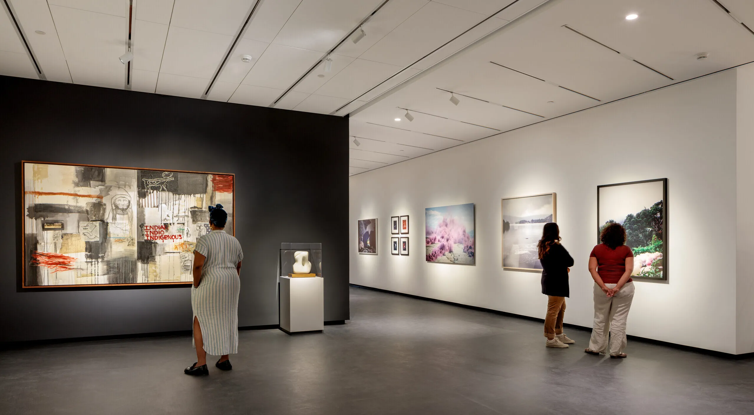 A modern museum gallery is photographed at a wide angle. Visitors observe large artworks on the walls.