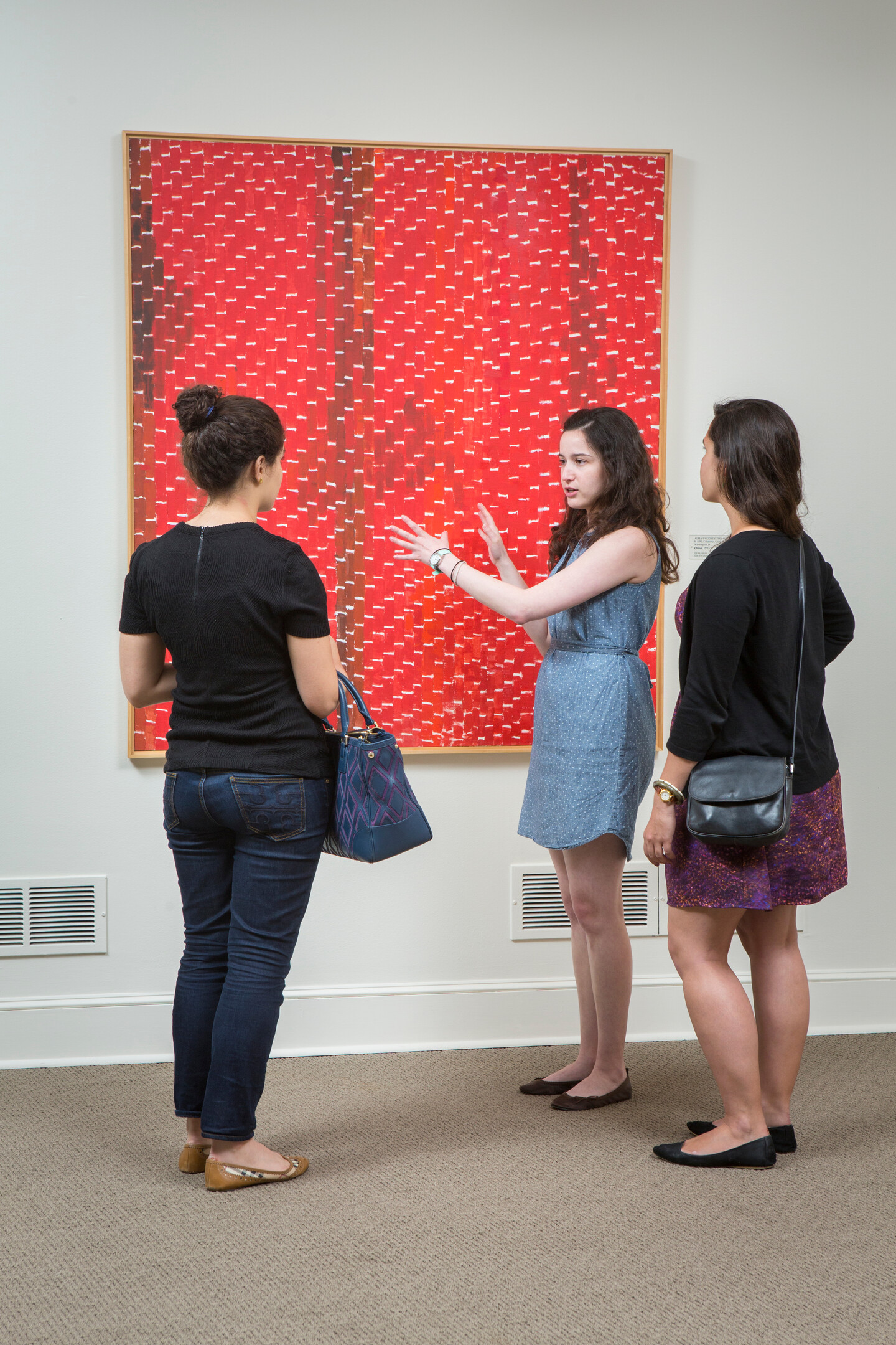 A woman with light skin tone and long dark hair in a denim summer dress talks about a large red and white abstract painting motioning with her arms outstretched. She is talking in front of two women who are looking at the painting in a museum gallery.