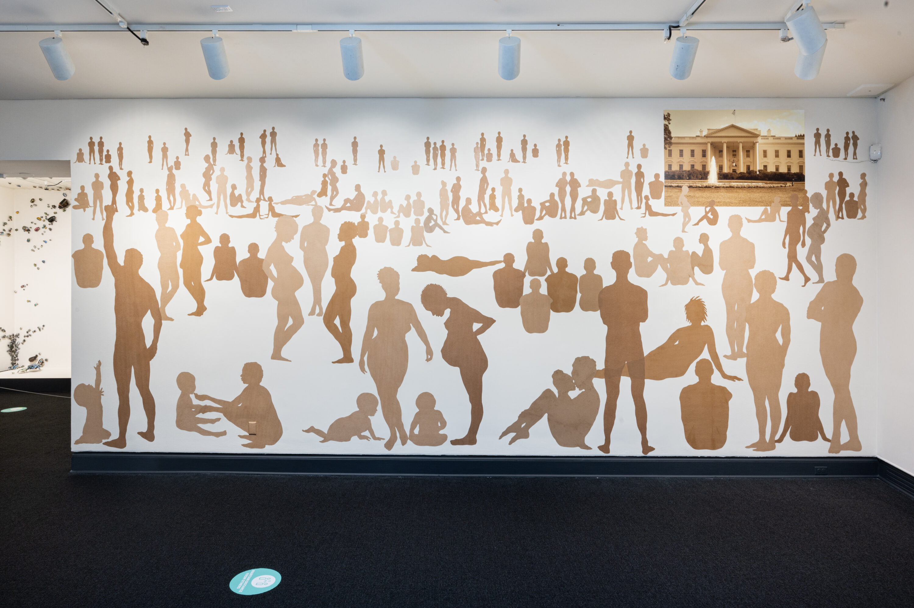 Over 100 silhouetted figures of varying ages, shapes, poses, and skin tones on a large white wall. In the top-right corner is a rectangular picture of the White House.