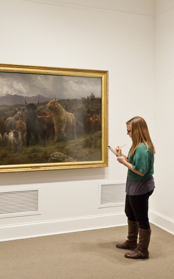 A young woman with light skin tone and long blonde hair wearing a green cardigan, gray shirt, black pants, and tall brown boots sketches with a notebook and pencil in front of a large painting of bulls and sheep.