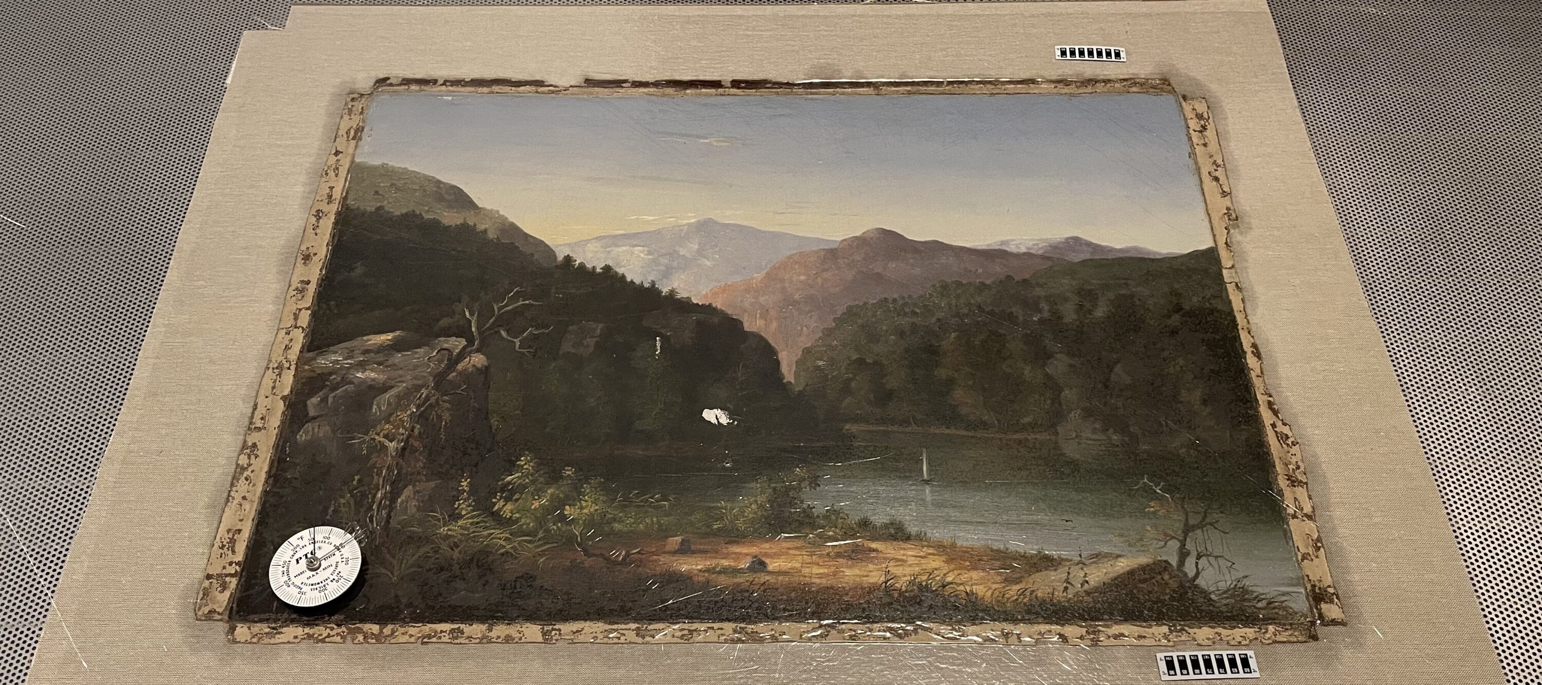 An oil painting of a mountainous landscape with a river cutting through it is laid on sterile table and prepared for conservation. A compass its in the lower left corner of the painting.