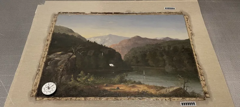 An oil painting of a mountainous landscape with a river cutting through it is laid on sterile table and prepared for conservation. A compass its in the lower left corner of the painting.