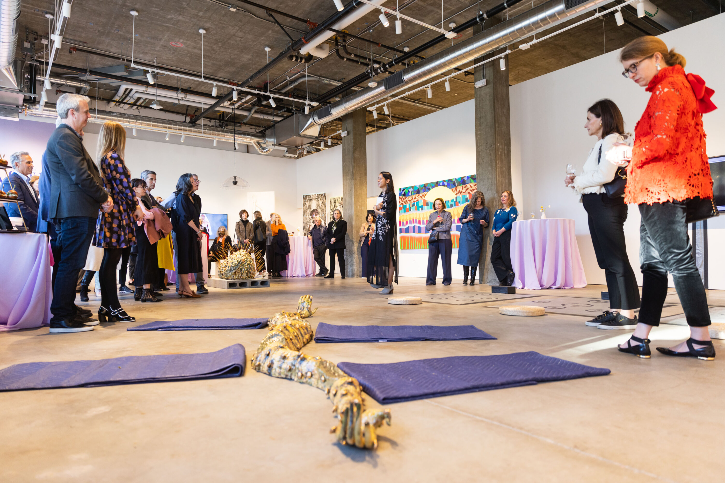 A large group attends an event in a gallery filled with colorful works on the walls and a large sculptural work in the middle of the floor. One figure stands in the middle of the room speaking to the group.