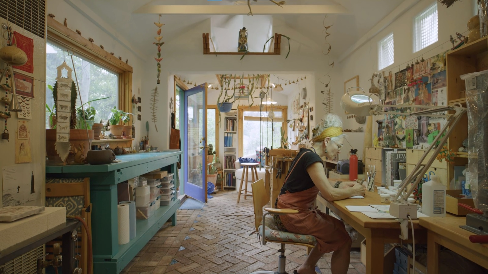 Film still of a woman with white hair sitting at a desk in the middle of an art studio filled with inspirational images on the walls and organic forms hanging from the ceiling.