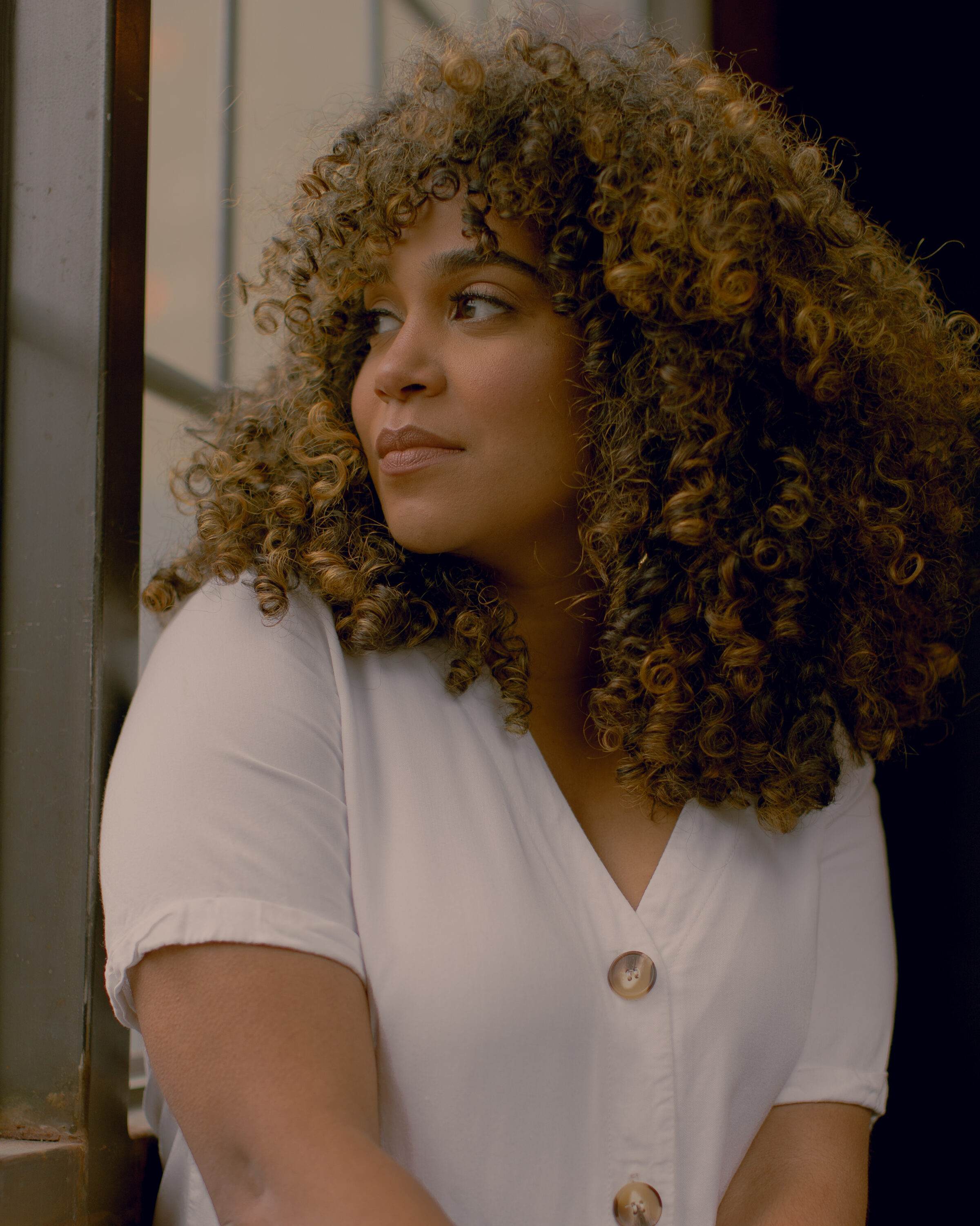 A dark-skinned woman with shoulder-length curly hair sits or stands in front of a window, looking over her shoulder and out of it. The light is somewhat dim. She wears a white button-up short sleeved shirt.