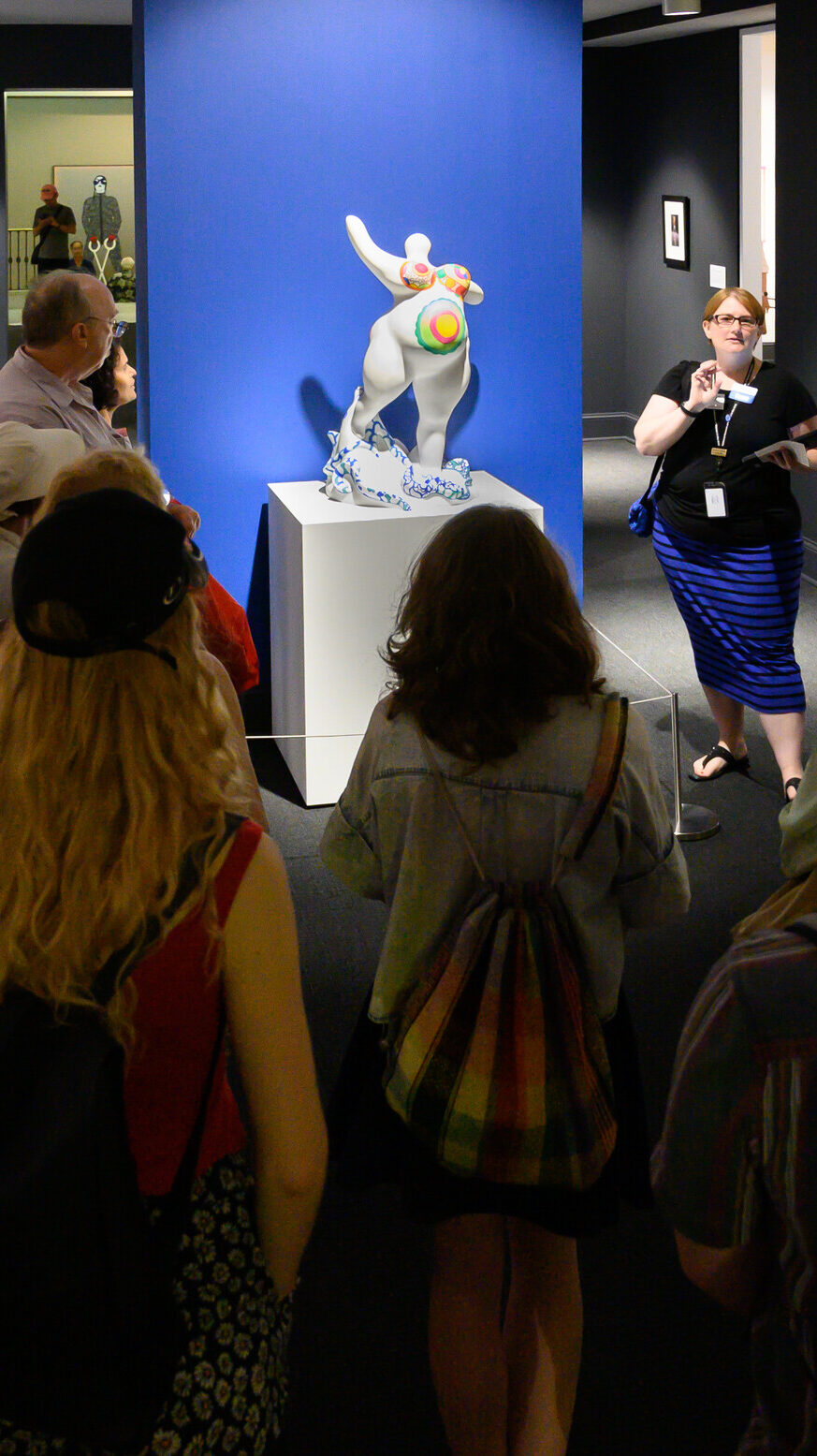 A light-skinned museum docent discusses a white marble sculpture of a female figure covered in bright patterns with visitors in the gallery.
