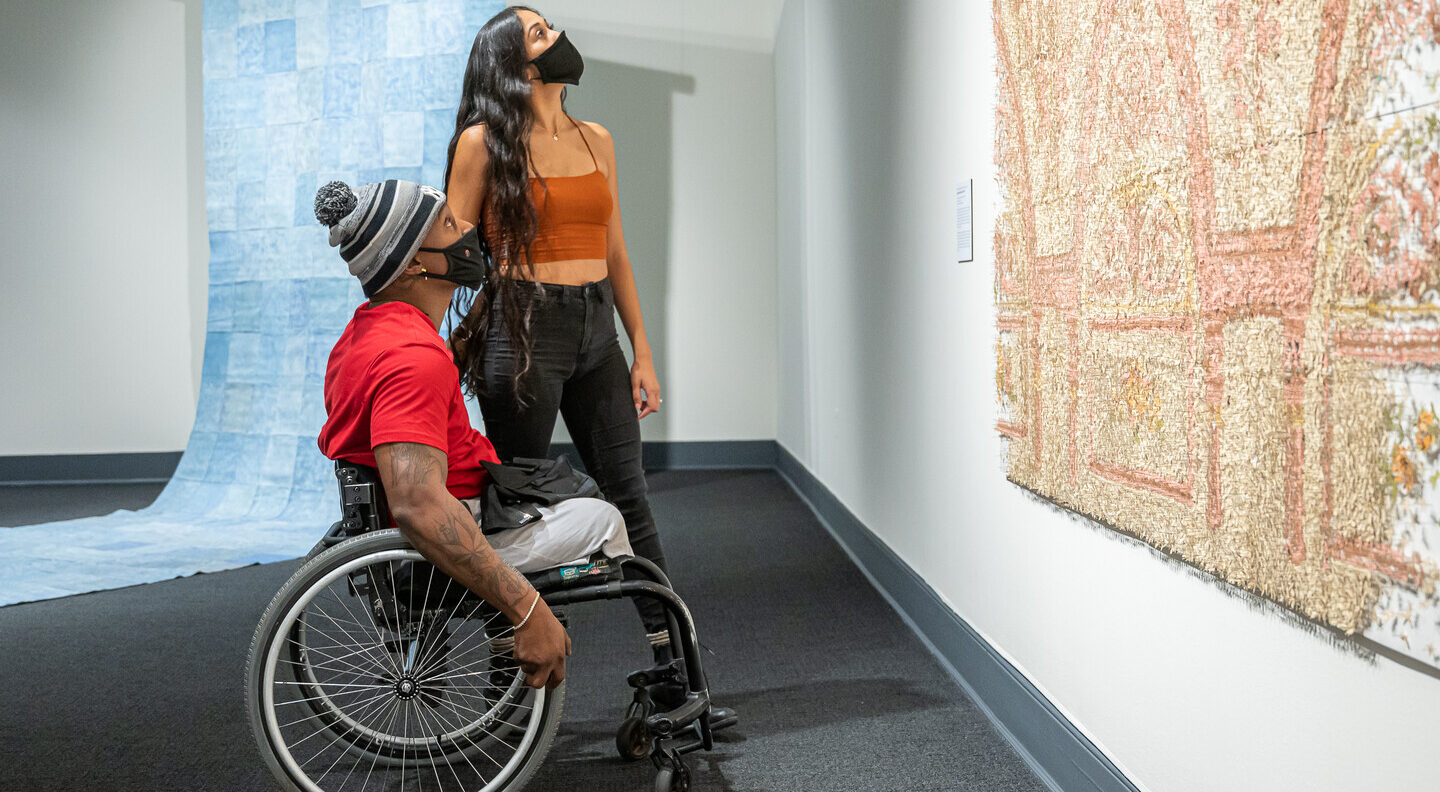 Two people look up at a large peach, tan, and ivory art installation in a gallery. The person with medium skin tone and tattoos on their right arm sits in a wheelchair and wears a red t-shirt, gray pants, gray-and-black striped winter hat, and black face mask. A person with medium skin tone, long curly black hair stands behind, wearing a burnt orange tank top, black jeans, and a black face mask. A large checkered blue artwork unfolds onto the gallery floor behind the pair.