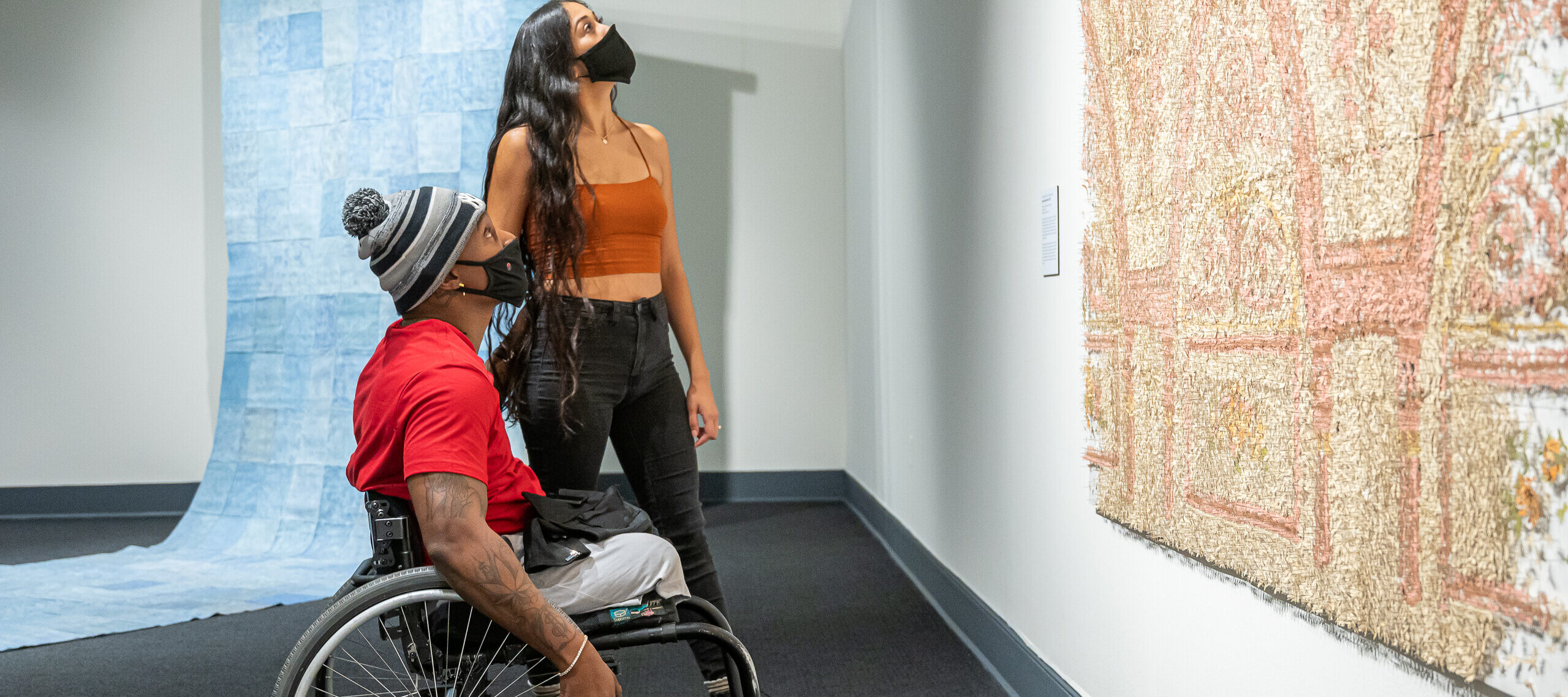 Two people look up at a large peach, tan, and ivory art installation in a gallery. The person with medium skin tone and tattoos on their right arm sits in a wheelchair and wears a red t-shirt, gray pants, gray-and-black striped winter hat, and black face mask. A person with medium skin tone, long curly black hair stands behind, wearing a burnt orange tank top, black jeans, and a black face mask. A large checkered blue artwork unfolds onto the gallery floor behind the pair.