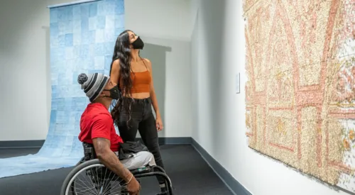 Two people look up at a large peach, tan, and ivory art installation in a gallery. The person with medium skin tone and tattoos on their right arm sits in a wheelchair and wears a red t-shirt, gray pants, gray-and-black striped winter hat, and black face mask. A person with medium skin tone, long curly black hair stands behind, wearing a burnt orange tank top, black jeans, and a black face mask. A large checkered blue artwork unfolds onto the gallery floor behind the pair.