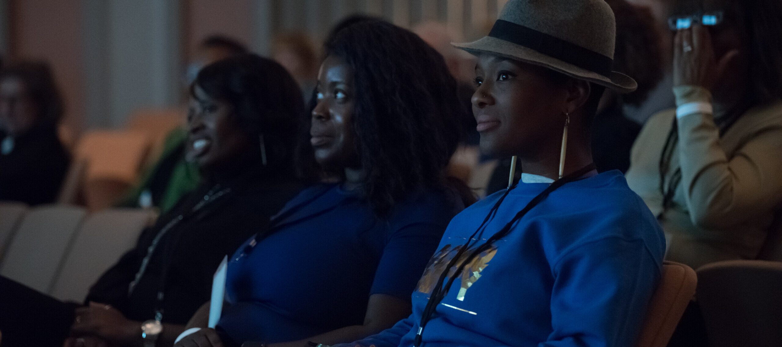 Three dark-skinned women sit in an auditorium with their hands crossed in their laps, intently watching an event.