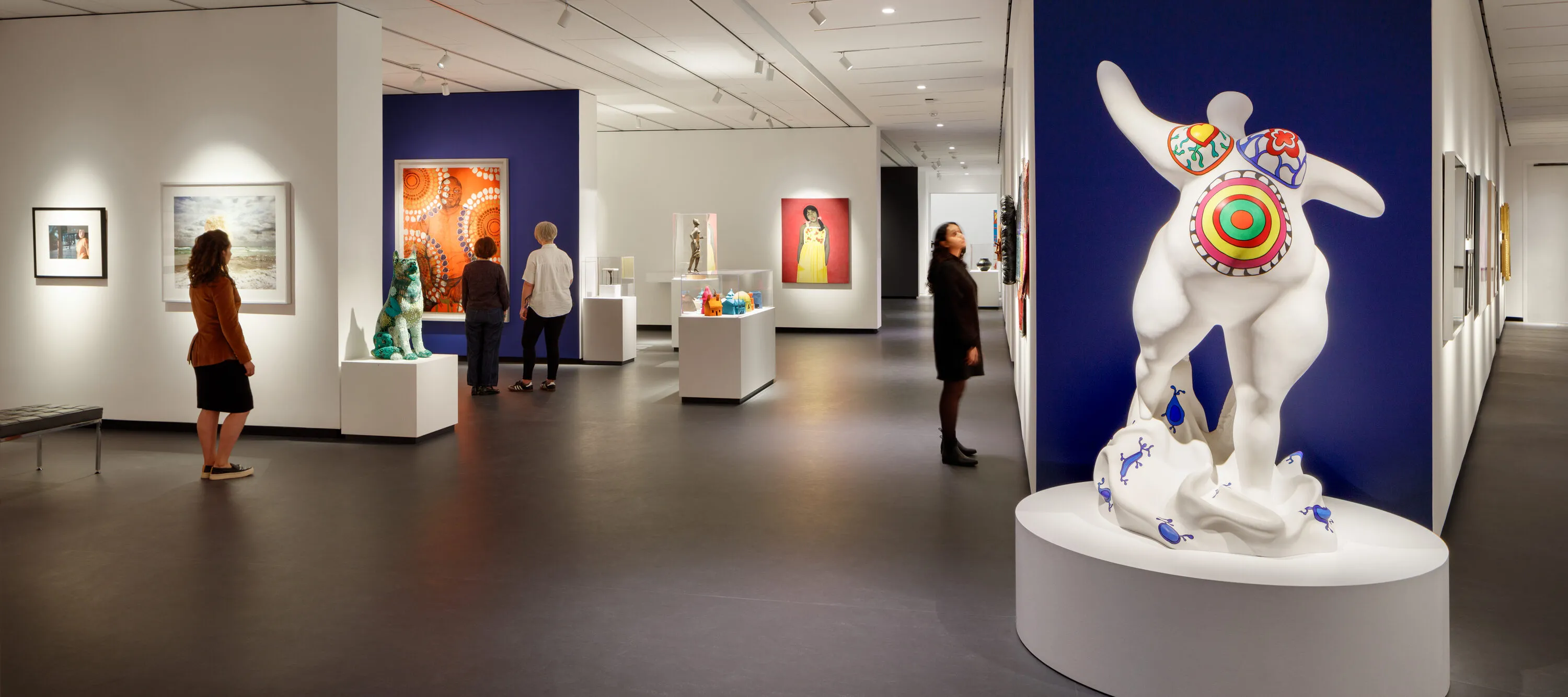 A modern museum gallery is photographed at a wide angle. It features several inset bays in which art of various sizes and mediums is hung. In the foreground, a marble sculpture depicts an abstracted, voluptuous figure with a pregnant belly. The figure is covered in bright patterns and posed stepping forward, with raised, outstretched arms.