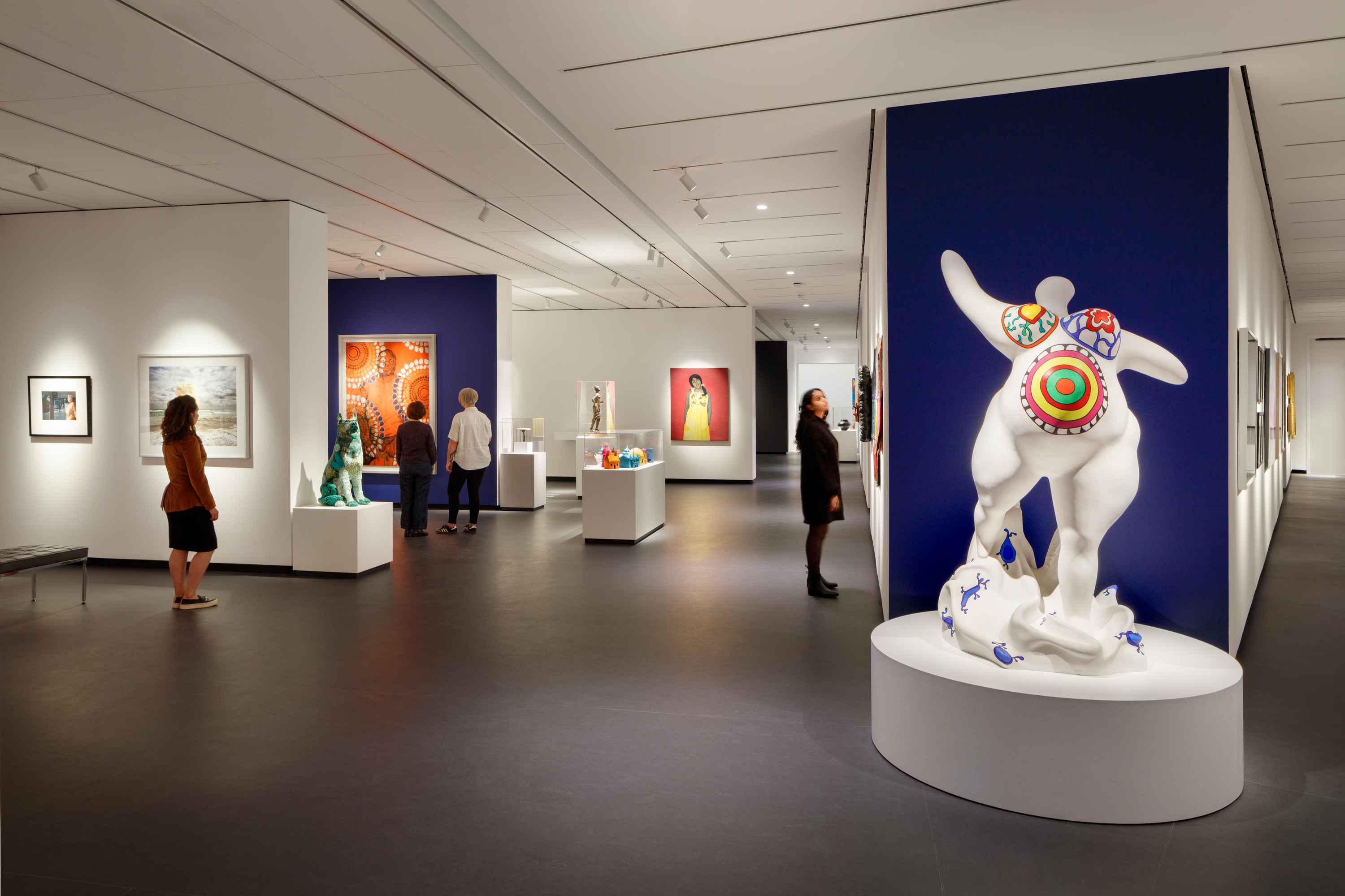 A modern museum gallery is photographed at a wide angle. It features several inset bays in which art of various sizes and mediums is hung. In the foreground, a marble sculpture depicts an abstracted, voluptuous figure with a pregnant belly. The figure is covered in bright patterns and posed stepping forward, with raised, outstretched arms.