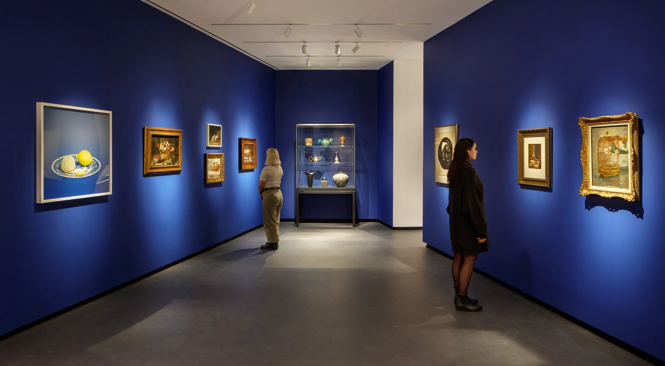 Museum visitors observe artworks hanging on the royal blue walls of a small gallery bay. Paintings of various sizes are on display, and small sculptures are in a glass case.