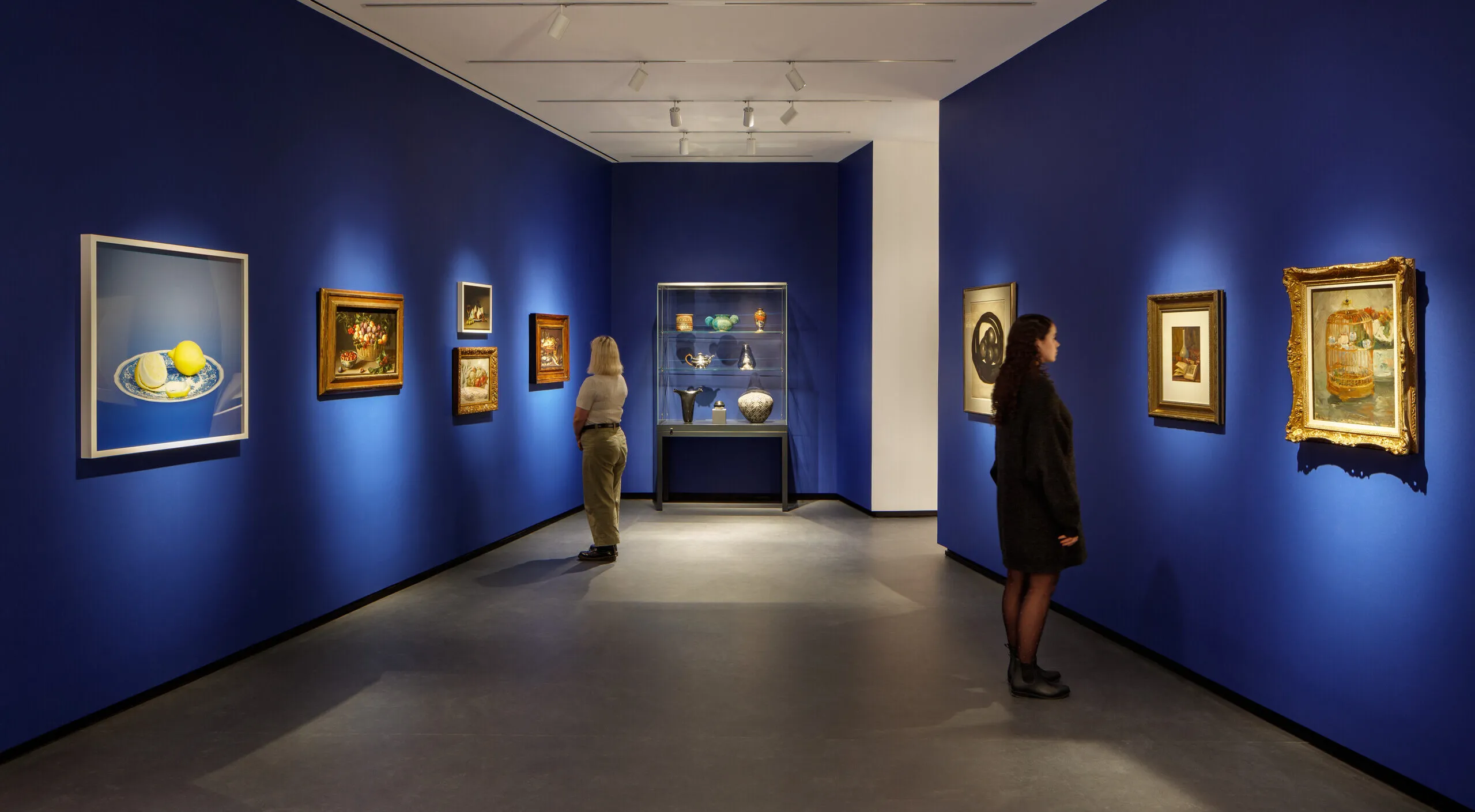 Museum visitors observe artworks hanging on the royal blue walls of a small gallery bay. Paintings of various sizes are on display, and small sculptures are in a glass case.