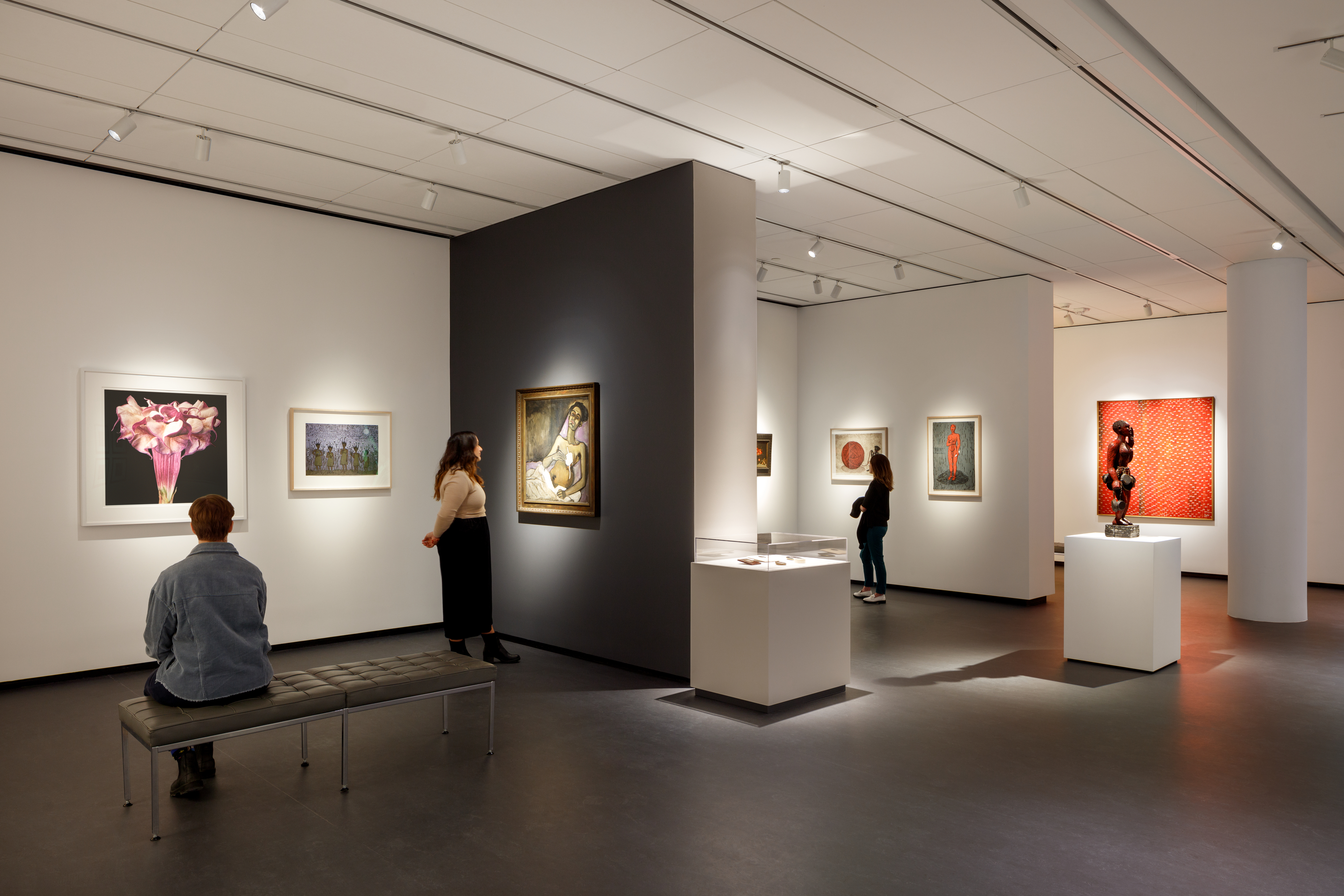 A modern museum gallery is photographed at a wide angle. It features several inset bays in which art of various sizes and mediums is hung. Visitors walk through the galleries and observe the works.