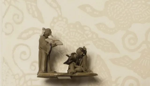 A woman wearing an elaborate hairstyle and earrings is seen in profile against a cream background and outlines of butterflies. A decorative glass bottle and two miniature ceramic sculptures of male figures are attached to the surface of the print.
