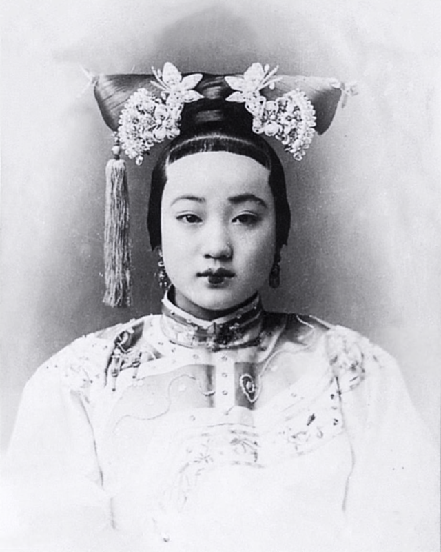 A black-and-white photograph shows a young Asian woman wearing traditional Chinese attire and an ornamented, tasseled headdress.