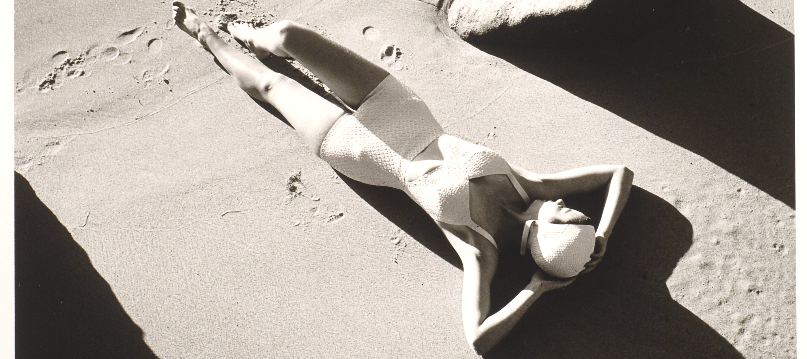 A black and white photograph of a light-skinned woman wearing a one-piece white bathing suit and white swim cap. She is lying down on sand with her hands behind her head. There are footprints in the sand below her, and the image is framed by large rocks.