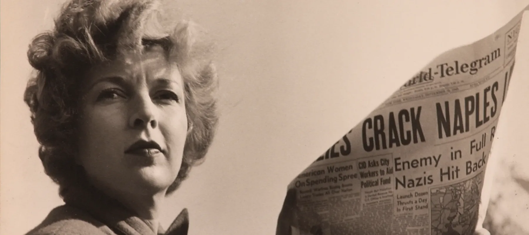 A black-and-white photograph of a light-skinned adult woman holding a newspaper with news about World War II. She wears a coat and her short, curly hair is caught in the wind.