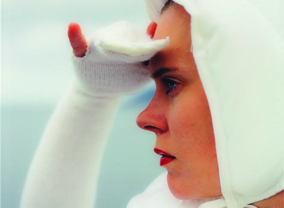 Square close-up of a light-skinned woman in profile looking to the left, seen from the shoulders up. She wears all white, including a bonnet and knit sleeves that become gloves covering her three middle fingers, and she holds her right hand up as if shielding her eyes from the sun.