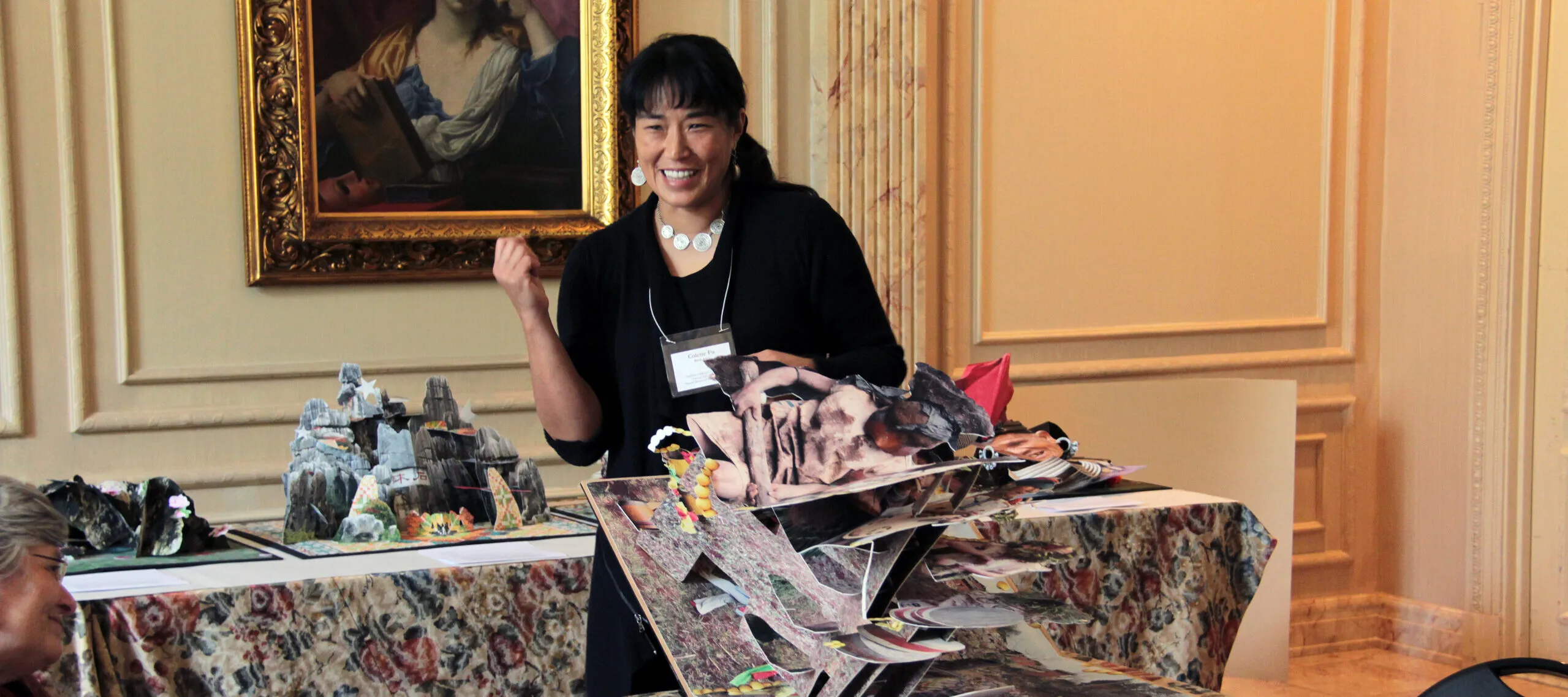 A woman with medium skin tone and dark hair wearing a black dress holds a large pop-up artist book, which unfolds in her hand and onto a table.