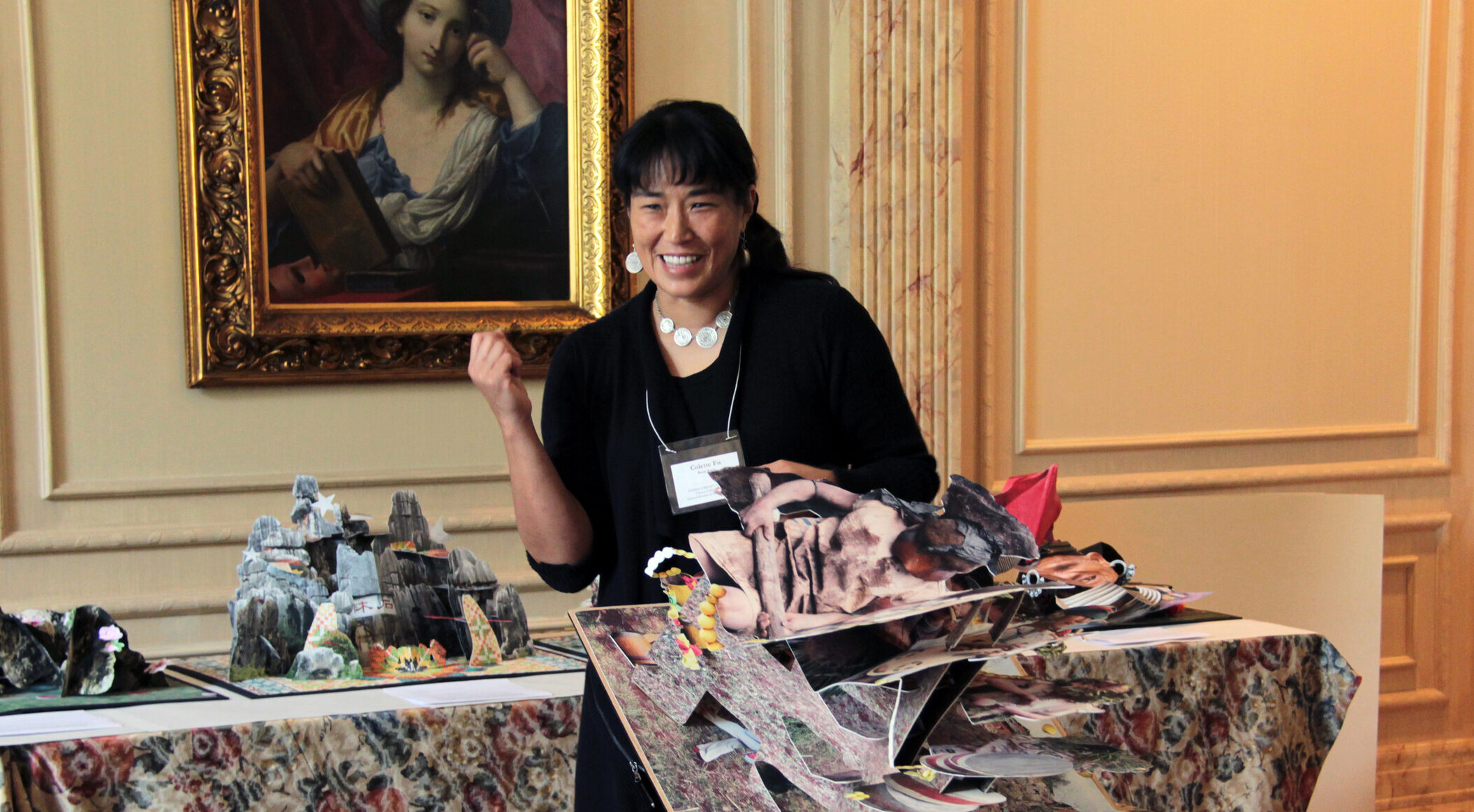 A woman with medium skin tone and dark hair wearing a black dress holds a large pop-up artist book, which unfolds in her hand and onto a table.