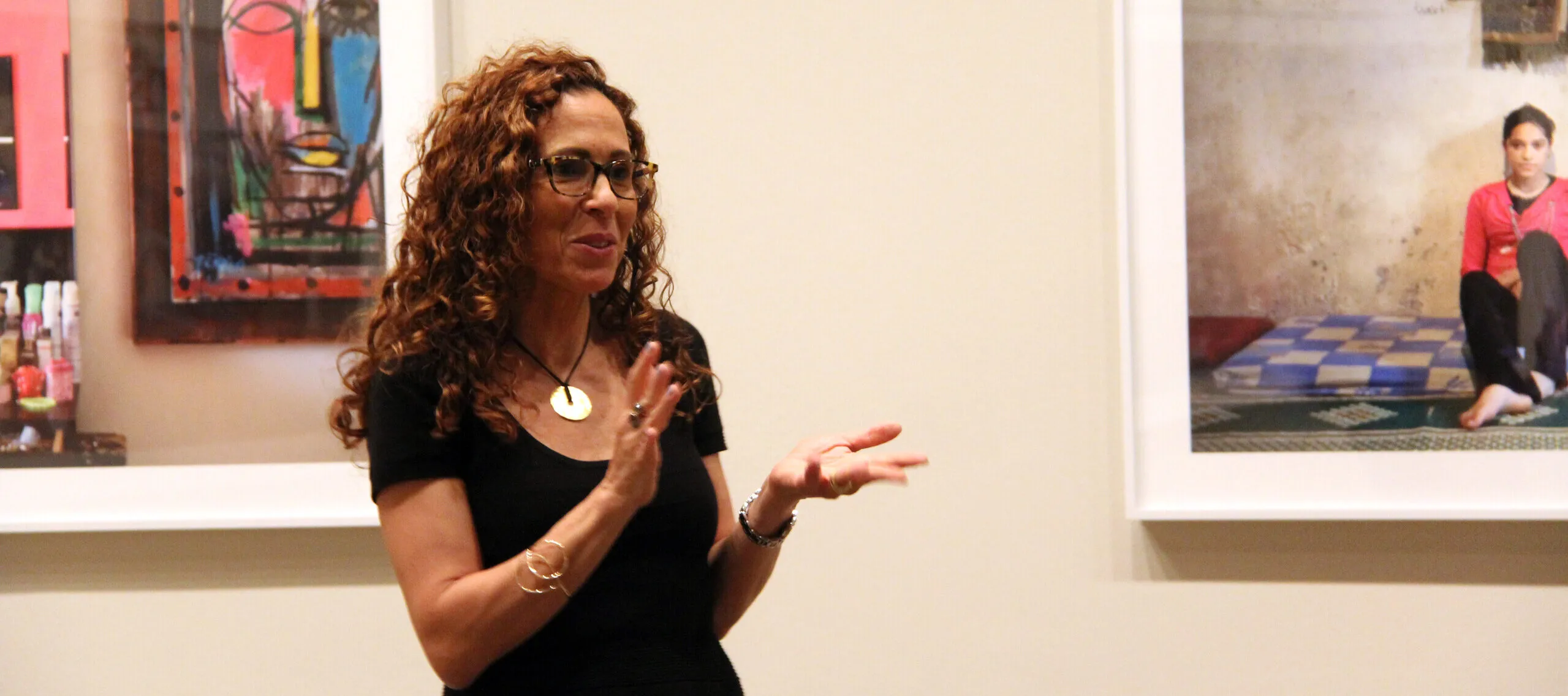 A woman with medium skin tone and curly auburn hair and glasses gestures with her hands as she speaks in a gallery between two colorful photographs.