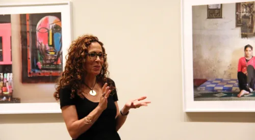 A woman with medium skin tone and curly auburn hair and glasses gestures with her hands as she speaks in a gallery between two colorful photographs.