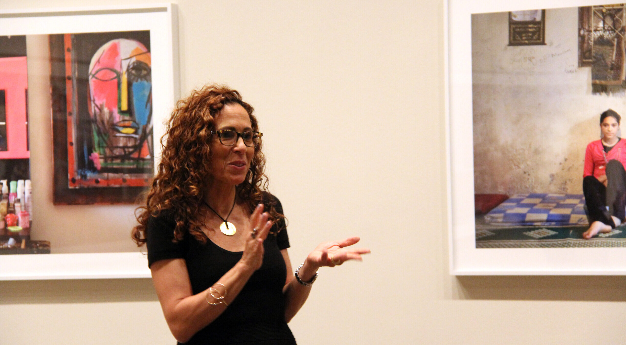A woman with medium skin tone and curly auburn hair and glasses gestures with her hands as she speaks in a gallery between two colorful photographs.