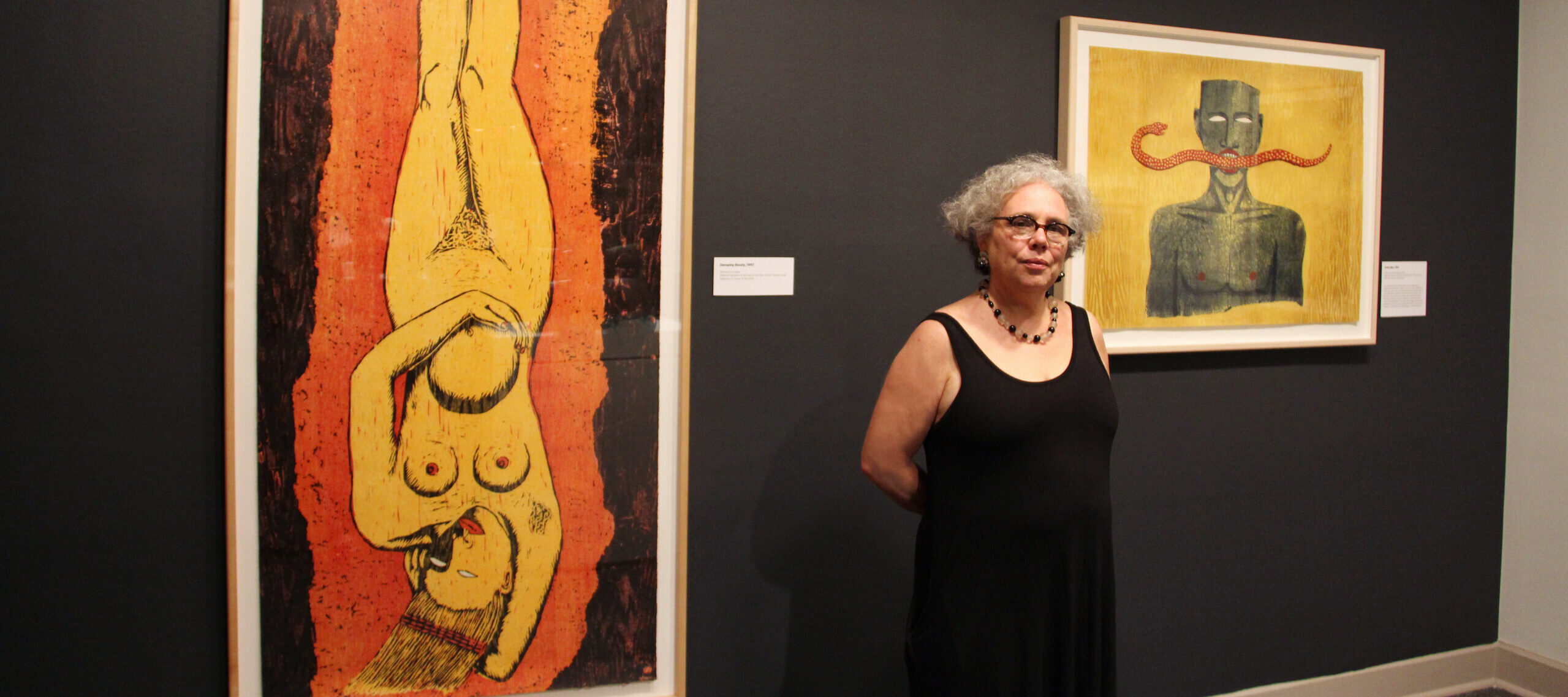 The artist, a woman with light skin, curly gray hair, and glasses, wears a black sleeveless dress and necklace while standing in a gallery in front of two of her large colorful prints.
