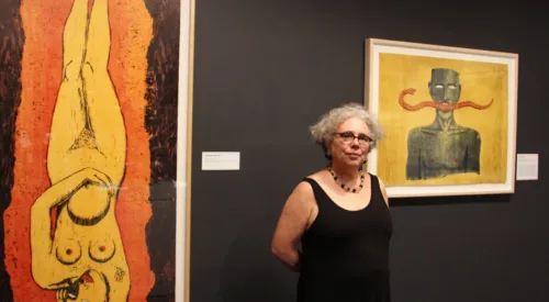 The artist, a woman with light skin, curly gray hair, and glasses, wears a black sleeveless dress and necklace while standing in a gallery in front of two of her large colorful prints.